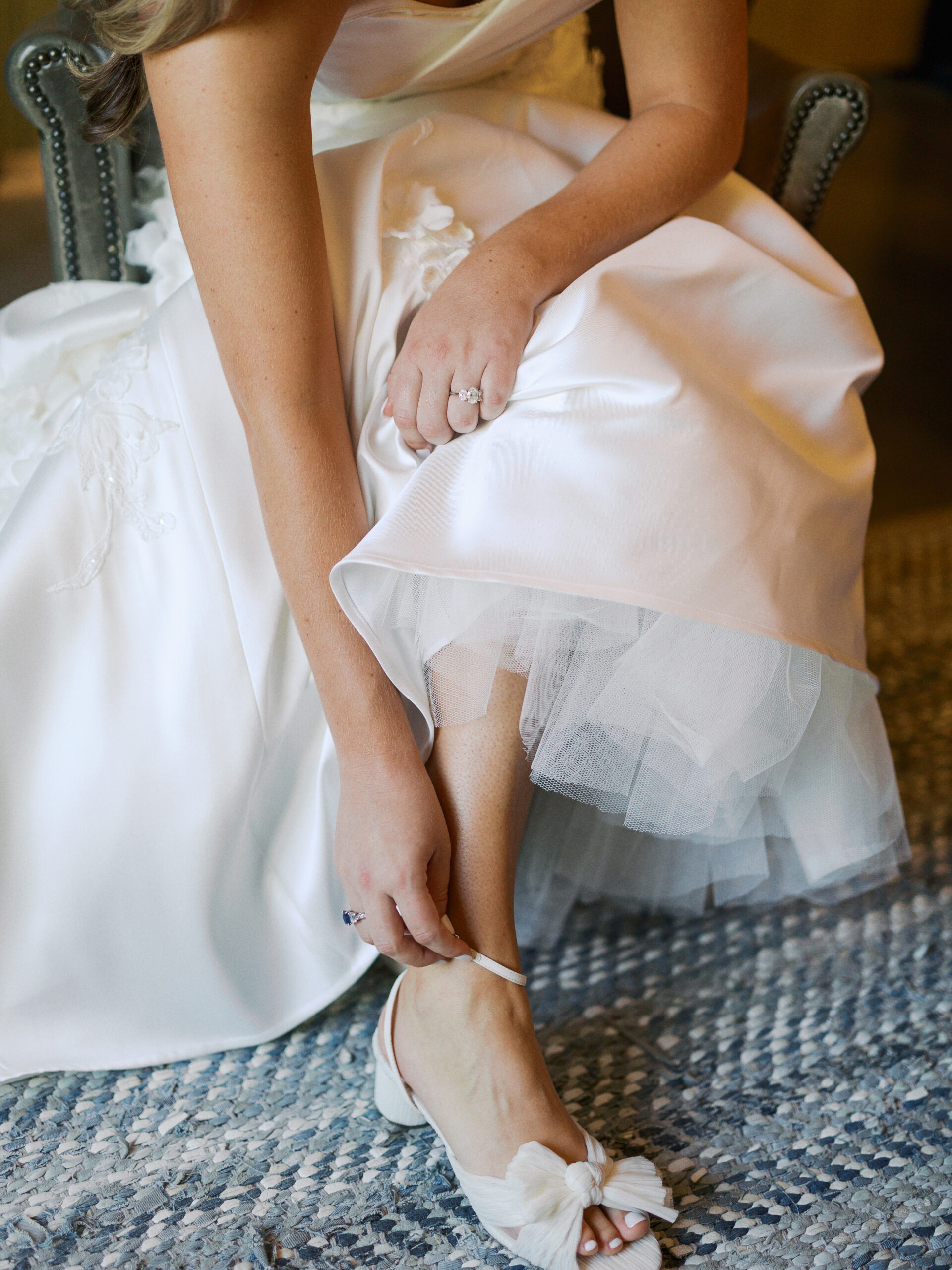Close-up of a bride in a white wedding gown sitting and fastening the strap of her elegant white wedding shoe.