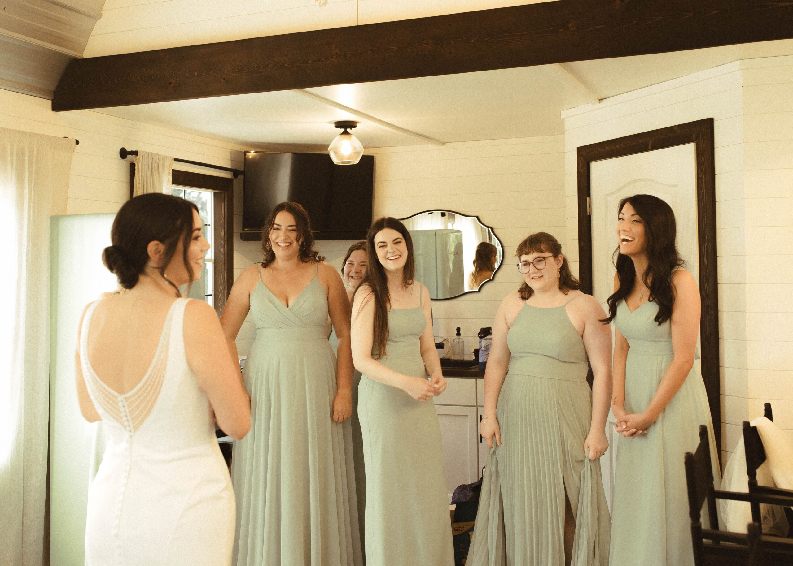 Joyful bride sharing a laugh with her smiling bridesmaids in sage green dresses during wedding preparations by Olive & Eden.