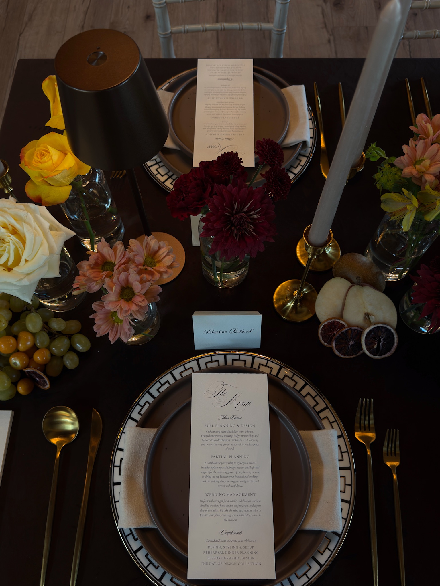 Vertical overhead shot of a moody luxury wedding tablescape featuring chocolate brown velvet linens, gold geometric chargers, and fruit accents by Austin wedding designer Olive & Eden.
