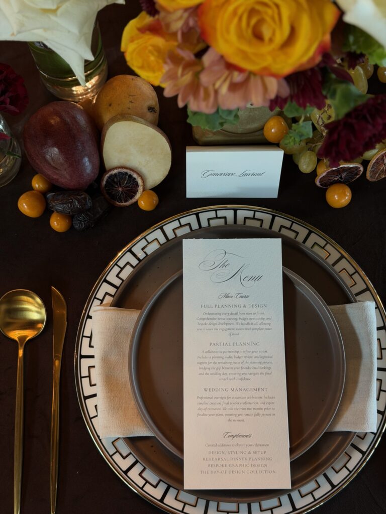 Top-down view of bespoke wedding menu card on brown matte plate with gold geometric charger and fruit accents.