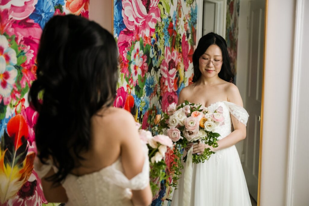 A bride reflecting in a mirror against a vibrant floral wall during getting ready moments at Hummingbird House.