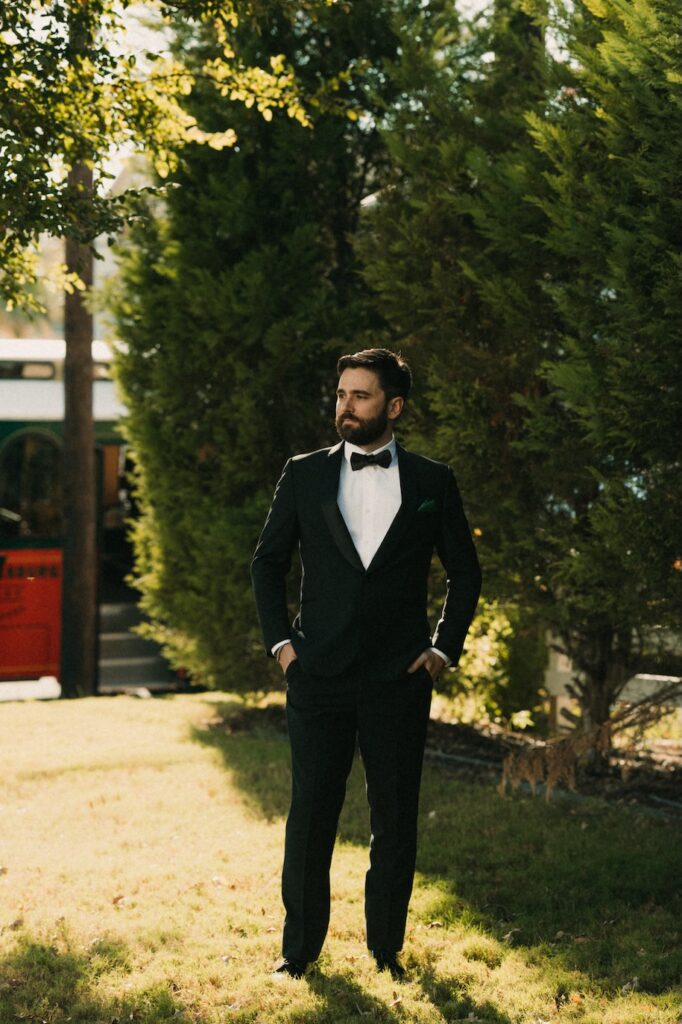 A modern groom in a black tuxedo standing outside near a vintage trolley in the Texas Hill Country.