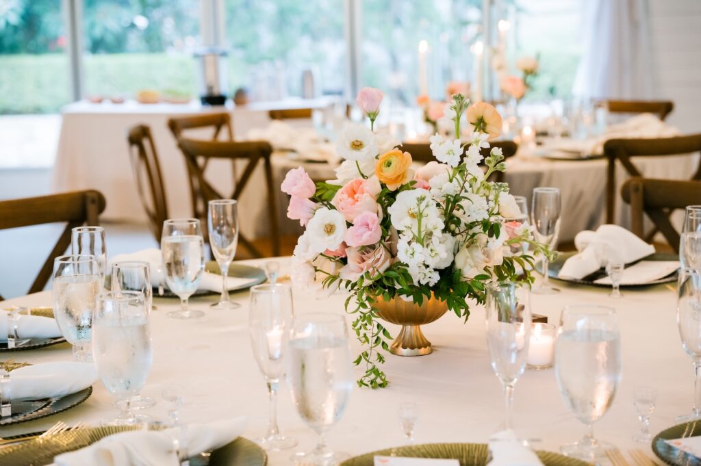 Elegant guest table setting featuring a gold compote floral arrangement, glassware, and glowing candlelight in a tented reception.