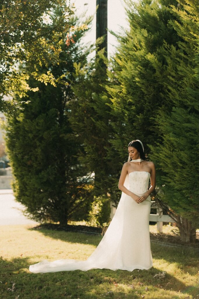 A full-length bridal portrait of a modern bride in a corset dress standing against lush evergreen trees in Fredericksburg.